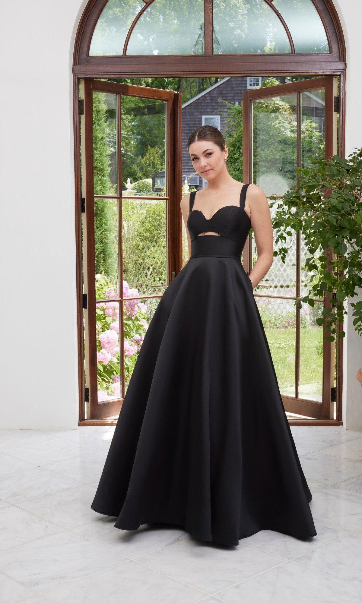 A woman wearing the Long Prom Dress: Morilee 49111 in black, featuring a sleeveless design, sweetheart neckline, and waist cutout, stands indoors by open glass doors with greenery visible outside.